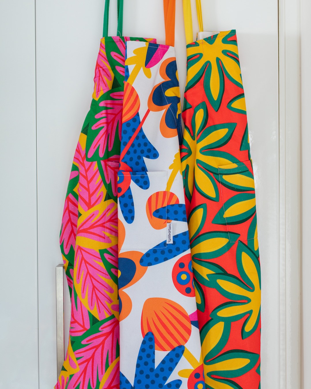 Three bold, colourful aprons with graphic prints hanging on a kitchen cupboard door